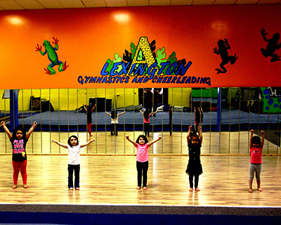 Five children with arms raised stand in a gym studio in front of a mirrored wall; orange wall behind them displays the 'Lexington Gymnastics and Cheerleading' logo.