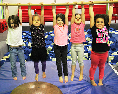 Five smiling girls hold onto a yellow bar and jump on a trampoline in a gym, surrounded by blue foam pit mats.