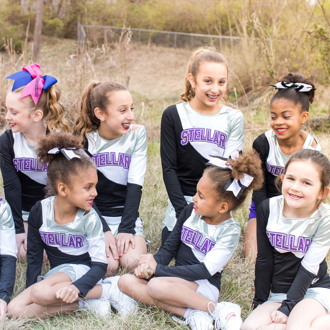 Group of diverse young cheerleaders in silver and black Stellar uniforms sitting in a grassy field, smiling and talking outdoors.