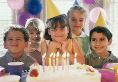 Kids wearing party hats smile around a frosted birthday cake with lit candles at a party hall.