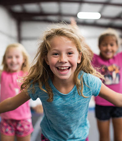 Center-front girl in a blue T-shirt laughs toward the camera while two teammates stand behind her in a gym class scene.