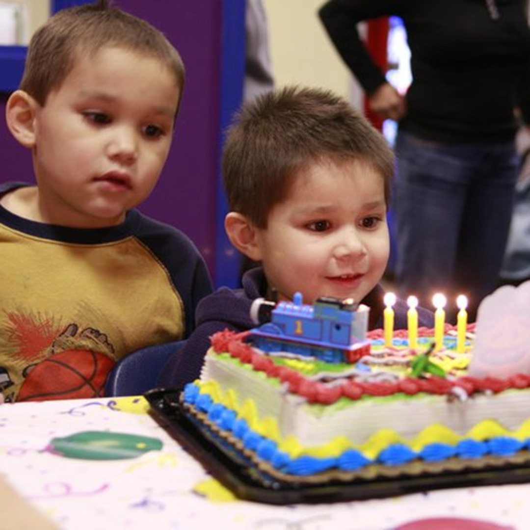 Two young boys watch a colorful train-themed birthday cake with lit candles.