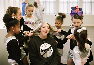 Smiling girl in a NASA sweatshirt sits center as six teammates style her hair backstage