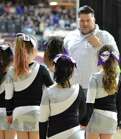 Young cheerleaders in silver and black uniforms with purple bows stand with backs to the camera as a coach speaks to them in a gym.