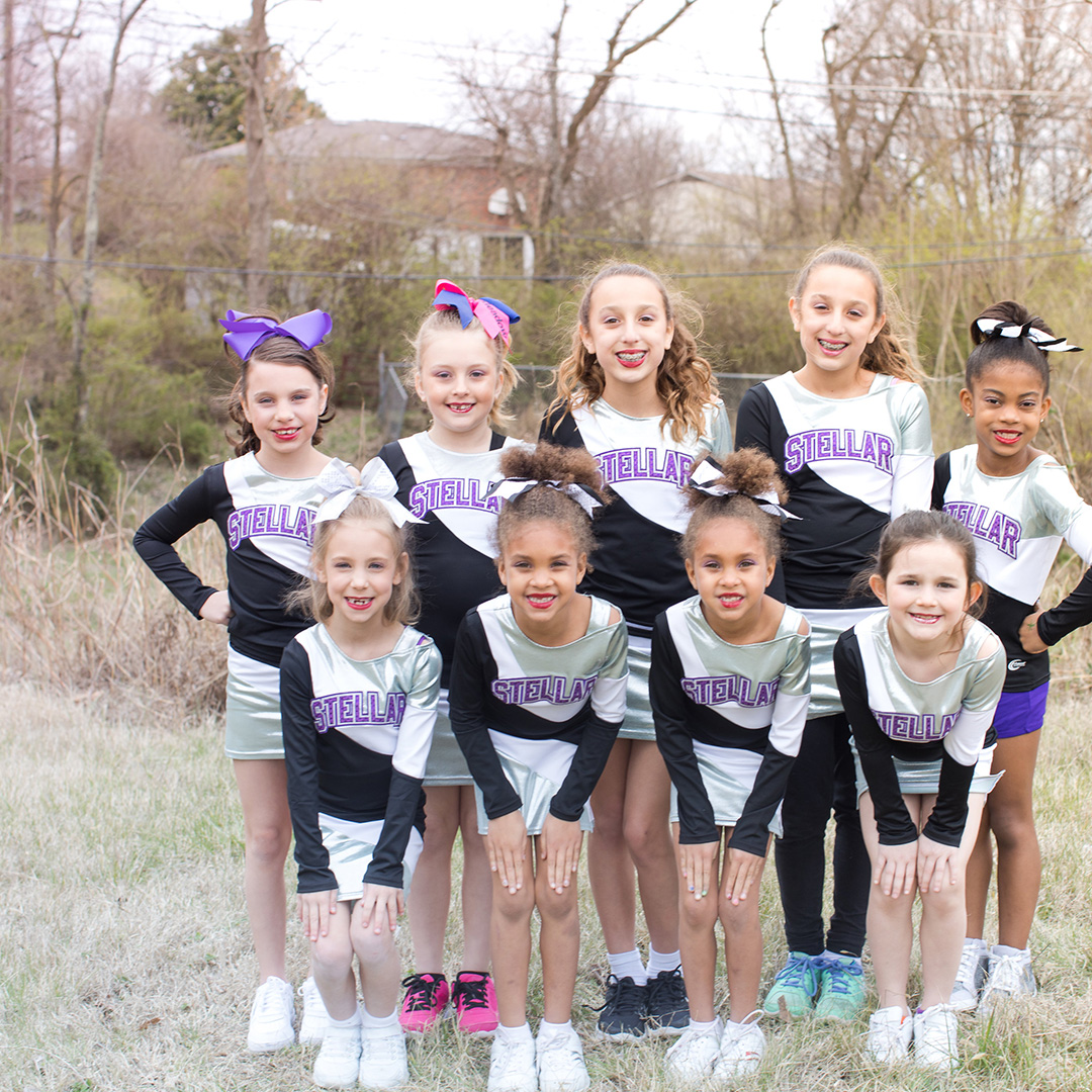 Group of ten young cheerleaders in silver and black uniforms with 'STELLAR' on their chests, posing outdoors in a grassy area with trees behind them.