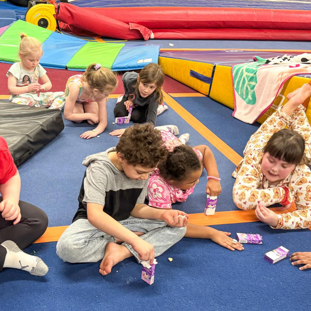 Group of children sitting on a blue gym mat, opening purple juice cartons during a class activity on colorful padded mats.