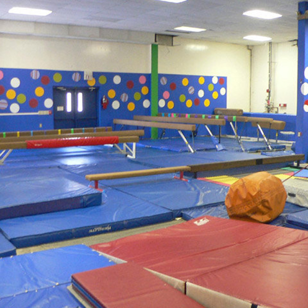 A gymnastics gym with blue mats, balance beams, and a padded spring floor in a colorful wall-lined room.