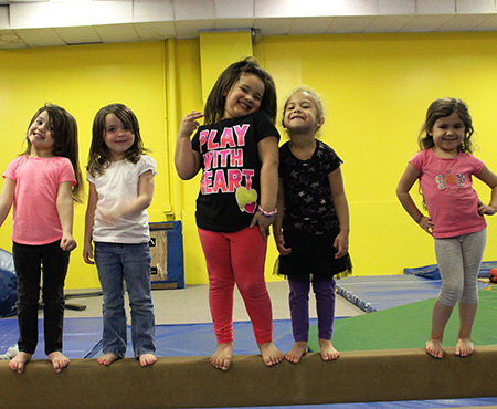 Five barefoot girls pose on a gym balance beam in a yellow-walled room, smiling at the camera while standing in a line.