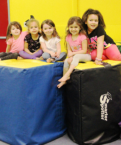 Five smiling young girls sit together on padded blue and black foam blocks in a bright indoor play area.