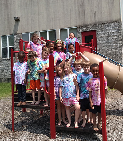 Group of children in tie-dye shirts standing on a playground platform and slide, smiling at the camera outdoors on a sunny day.