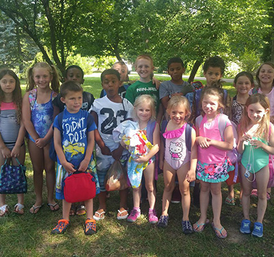 Group of diverse young children standing together on grass in a sunny park with trees in the background, wearing colorful outfits and carrying bags.