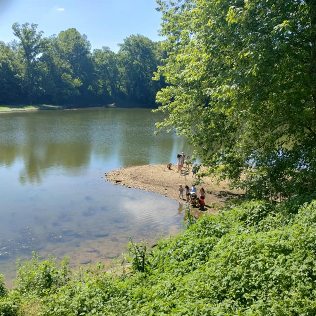 People enjoy a sunny day on a small sandy peninsula by a calm lake, framed by green trees.